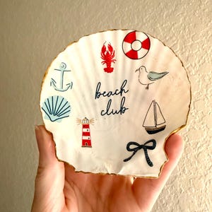 May include: A white scallop shell dish with a gold-painted rim, featuring the text "beach club" in black script. The shell is decorated with small illustrations of a red lobster, a red and white lifebuoy, a seagull, a sailboat, a lighthouse, an anchor, a blue shell, and a black rope knot.