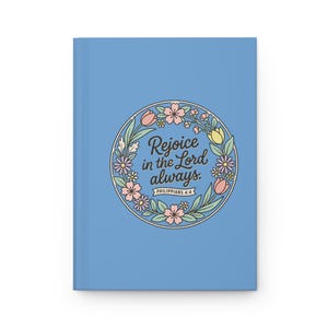 May include: A light blue journal featuring a floral wreath design. The wreath encircles the text "Rejoice in the Lord always" and "Philippians 4:4". The journal has a simple, minimalist aesthetic.