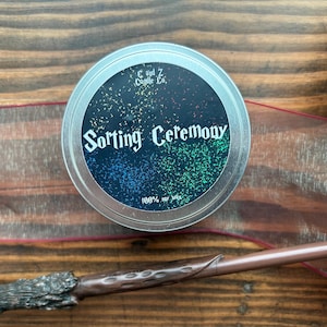 May include: A silver tin candle with a black label that reads "Sorting Ceremony" in white script. The label has a colorful speckled design. A wand, ribbon, and book are also visible on a wooden surface.