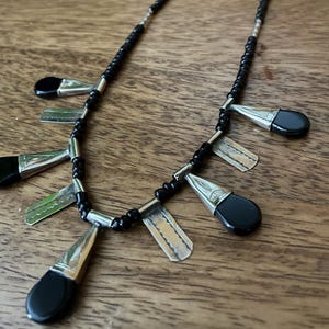 May include: A black beaded necklace with silver accents and teardrop-shaped black pendants. The necklace features a series of small black beads and silver-colored metal elements. The pendants are a combination of silver metal and black stone.