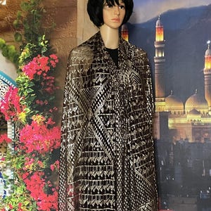 Puede incluir: Un chal negro y dorado con un diseño geométrico y detalles dorados metálicos, drapeado sobre un maniquí. El fondo incluye un mural de una ciudad de Oriente Medio con mezquitas y minaretes, y un arreglo floral.