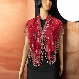 May include: A red, sheer shawl with a geometric pattern and beaded fringe. The shawl is draped over a black top. The design features small white accents and a delicate, open weave.