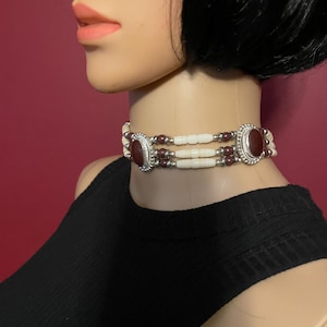 May include: A choker necklace with three strands. The top and bottom strands feature white beads, while the middle strand has small, dark red beads. The necklace has two large, oval-shaped, dark red stones with silver-colored metal settings.