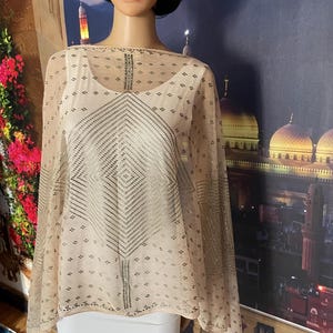 May include: A sheer, beige cape with a geometric silver pattern and small silver accents. The cape is draped over a white top and a white skirt. The cape has a loose, flowing design.