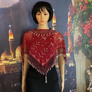 May include: A red shawl with intricate silver beadwork and a delicate, open-weave design. The shawl is draped over a mannequin, showcasing its triangular shape and beaded fringe. The background features a scenic backdrop.