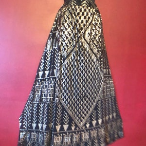 Puede incluir: Una falda larga negra con un intrincado patrón geométrico dorado. La falda se exhibe en un maniquí sobre un fondo rojo. El diseño incluye formas de diamante, cuadradas y de flecha, creando una apariencia texturizada.