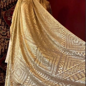 Puede incluir: Un chal metálico dorado con un patrón geométrico. El chal está hecho de una tela suave y transparente y tiene un diseño delicado e intrincado.