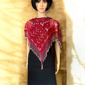 May include: A burgundy shawl with a geometric pattern and beaded fringe. The sheer fabric features a repeating design in white. The shawl is draped over a black dress on a mannequin, showcasing its triangular shape and decorative elements.