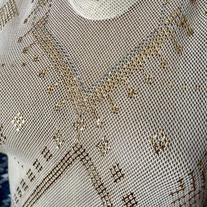 May include: Off-white mesh top with gold and silver embellishments. The top features a crew neck and intricate geometric patterns. A pearl necklace is visible at the neckline. The fabric has a delicate, open weave.