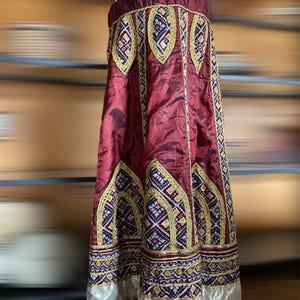 May include: A long, burgundy skirt with intricate embroidery and gold trim. The skirt features a repeating pattern of embroidered arches and geometric designs in white, yellow, and dark blue. The hemline is accented with silver fabric.