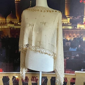 May include: A cream-colored shawl with gold embellishments and a delicate, fringed trim. The shawl is draped over a mannequin, showcasing its intricate design and lightweight fabric. The design includes small geometric patterns and gold beads.