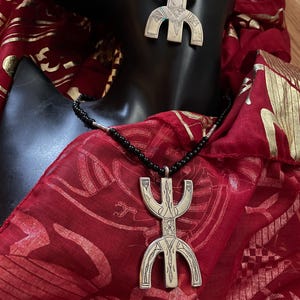 May include: A silver necklace and earrings with a traditional Ethiopian symbol pendant. The necklace is on a black cord and the earrings are hanging from a mannequin's ear. The jewelry is displayed on a red fabric with a gold pattern.