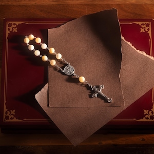 May include: A rosary with cream-coloured beads and a silver crucifix rests on a maroon book with gold trim. A torn piece of brown paper partially covers the book. The background is a wooden surface.