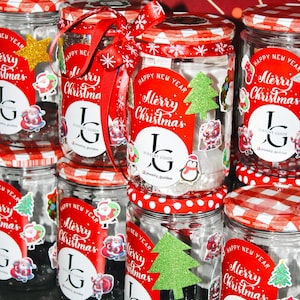 May include: A collection of glass jars with red and white chequered lids, decorated for Christmas. Each jar features a red label with "Merry Christmas" and festive stickers. Some jars are adorned with red ribbons and snowflakes, creating a festive display.