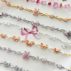 May include: Several delicate bracelets in gold and silver, featuring pink gemstones, floral designs, butterflies, and bows. The bracelets are displayed on a pink floral plate, highlighting their intricate details and craftsmanship.