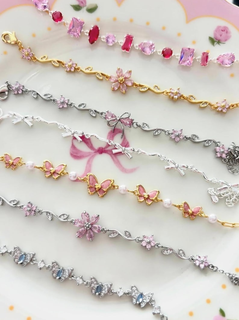 May include: Several delicate bracelets in gold and silver, featuring pink gemstones, floral designs, butterflies, and bows. The bracelets are displayed on a pink floral plate, highlighting their intricate details and craftsmanship.