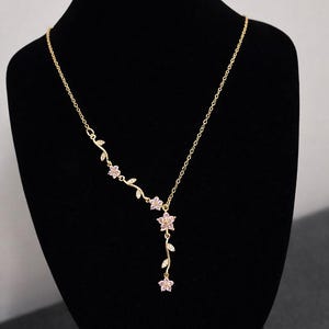 Può includere: Una collana color oro con un design floreale. La collana presenta una delicata catena con un motivo a vite adornato con piccoli fiori e foglie rosa. Il pendente è appeso, mettendo in mostra la composizione floreale.