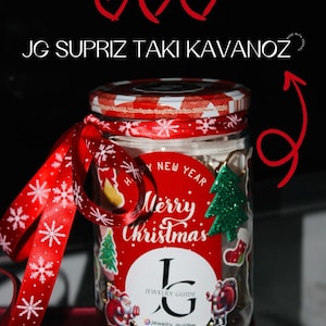 May include: A glass jar with a red and white checkered lid, adorned with a red ribbon and a Christmas-themed label. The label says "Merry Christmas" and features a Christmas tree and Santa Claus illustrations. The jar also has the text "JG SUPRIZ TAKI KAVANOZ".