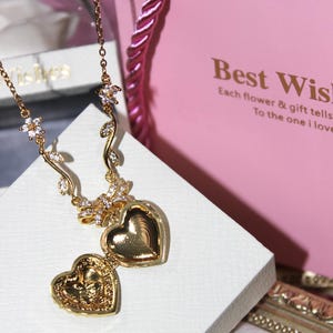 Puede incluir: Un collar dorado con un colgante en forma de corazón con detalles florales y de hojas. El relicario está abierto, revelando el interior. El collar se exhibe sobre una superficie blanca, con una bolsa de regalo rosa en el fondo.