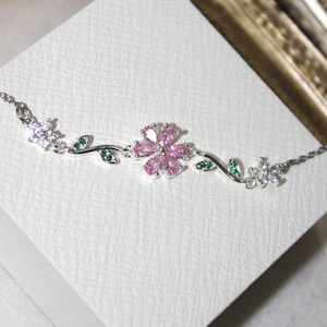Puede incluir: Una pulsera de plata con un diseño floral. La pulsera presenta una flor rosa central con piedras transparentes, hojas verdes y flores blancas más pequeñas. La pulsera se muestra sobre una superficie blanca texturizada.