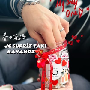 May include: A clear glass jar decorated with red hearts and stickers, tied with a red ribbon. The jar has text that reads "Happy Valentine's Day" and "Jewelry Guide". The jar is held in a person's hands.