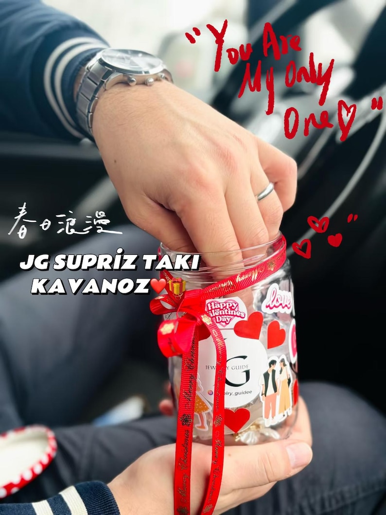 May include: A clear glass jar decorated with red hearts and stickers, tied with a red ribbon. The jar has text that reads "Happy Valentine's Day" and "Jewelry Guide". The jar is held in a person's hands.
