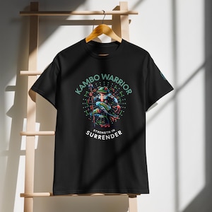 Könnte beinhalten: Schwarzes T-Shirt mit einer farbenfrohen Illustration eines Froschkriegers, der einen Stab hält. Der Text "KAMBO WARRIOR" wölbt sich über dem Frosch, mit "STRENGTH IN SURRENDER" darunter. Das Design hat grüne, blaue, rote und gelbe Akzente.