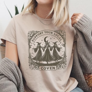 May include: Beige t-shirt featuring a graphic of three witches holding hands under a crescent moon and stars, with the text "SUPPORT YOUR LOCAL COVEN" in a decorative frame.