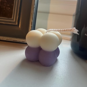 May include: A decorative candle in a unique bubble shape. The candle has a light purple base with white spheres on top. A natural-coloured wick extends from the top. The candle is on a white surface.