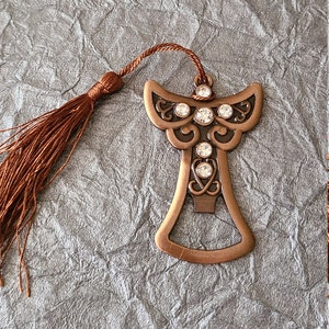Puede incluir: Un marcapáginas de ángel de color cobre con un borla marrón. El ángel tiene un diseño detallado y está adornado con diamantes de imitación transparentes.