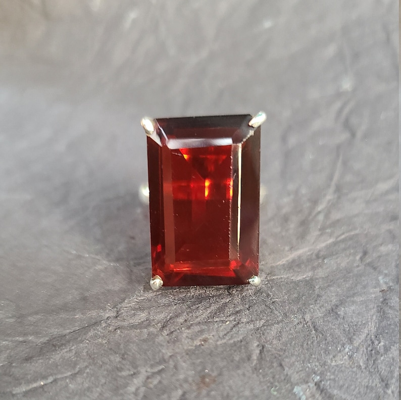 K&ouml;nnte beinhalten: Ein Ring mit einem rechteckigen, smaragdgeschliffenen roten Edelstein, der in einer silbernen Fassung sitzt.