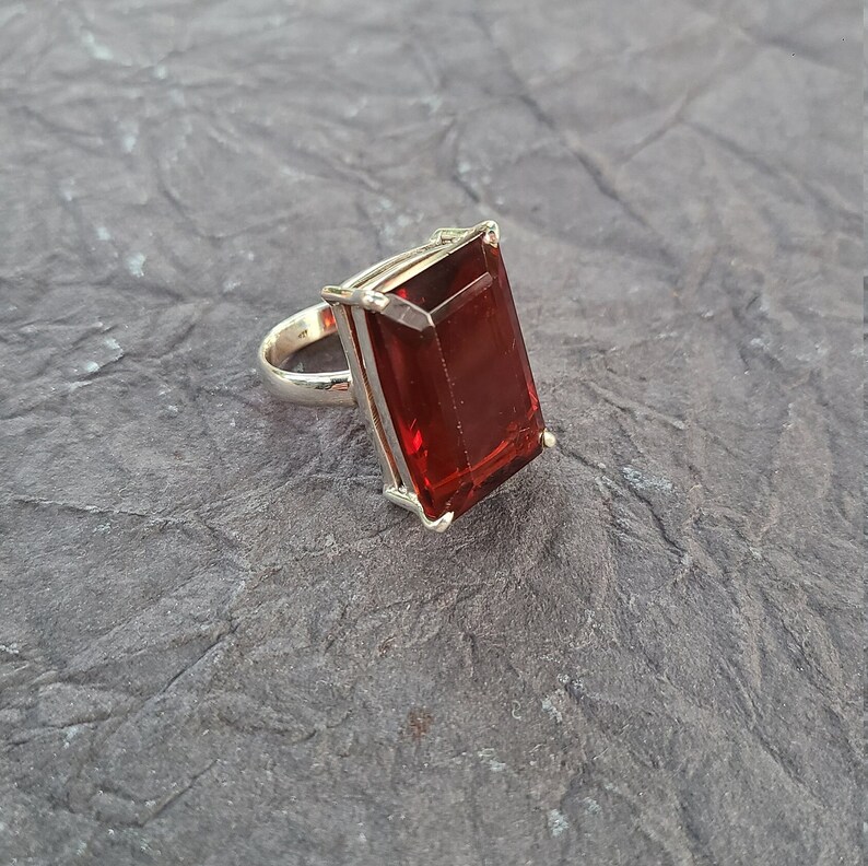 K&ouml;nnte beinhalten: Ein silberner Ring mit einem gro&szlig;en, rechteckigen, roten Edelstein. Der Edelstein ist in einer facettierten Form geschliffen.