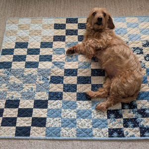 May include: A square quilted blanket featuring a patchwork design of alternating blue and cream fabric squares. The quilt displays various patterns including floral prints, stripes, and solid colors, finished with a light blue border and stitching.