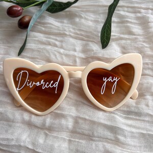 May include: White heart-shaped sunglasses with brown lenses. The left lens says "Divorced" and the right lens says "yay".