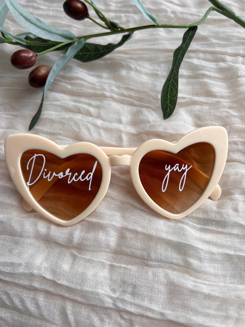 May include: White heart-shaped sunglasses with brown lenses. The left lens says "Divorced" and the right lens says "yay".