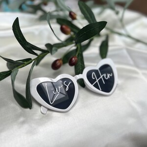 May include: White heart-shaped sunglasses with the text "Liv's Hen" written on them. The sunglasses are lying on a white surface with a green plant in the background.