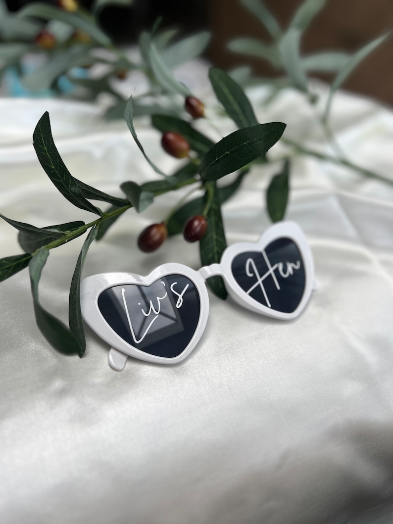 May include: White heart-shaped sunglasses with the text "Liv's Hen" written on them. The sunglasses are lying on a white surface with a green plant in the background.