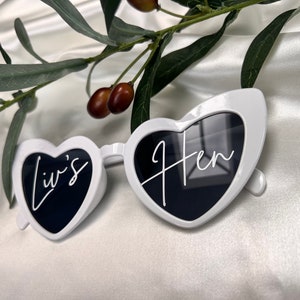 May include: White heart-shaped sunglasses with black lenses. The left lens says "Liv's" and the right lens says "Hen".