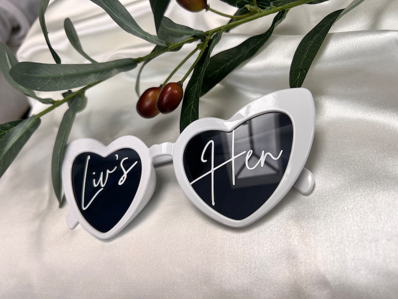 May include: White heart-shaped sunglasses with black lenses. The left lens says "Liv's" and the right lens says "Hen".