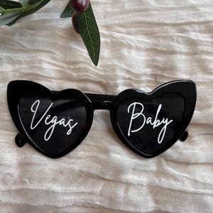 May include: Black heart-shaped sunglasses with white text. The left lens says "Vegas" and the right lens says "Baby".