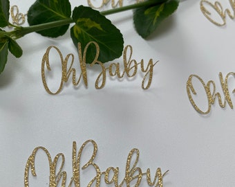 May include: Gold glitter confetti with the phrase "Oh Baby" written on each piece. The confetti is scattered on a white surface with a green plant stem.