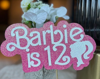 May include: Pink glitter card with white lettering that says "Barbie is 12" with a white glitter silhouette of a Barbie doll.