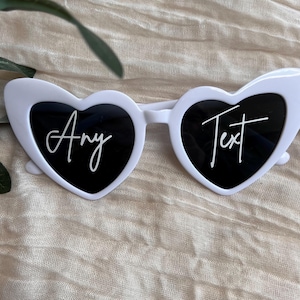 May include: White heart-shaped sunglasses with black lenses. The sunglasses have the text "Any Text" written on the lenses in white.