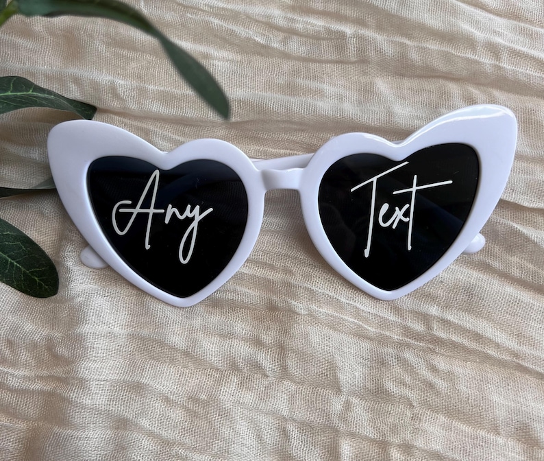 May include: White heart-shaped sunglasses with black lenses. The sunglasses have the text "Any Text" written on the lenses in white.