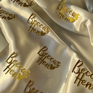 May include: Gold glitter 'Beccas Hen' party decorations on a white satin background.