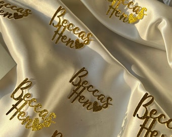 May include: Gold glitter 'Beccas Hen' party decorations on a white satin background.