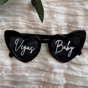 May include: Black heart-shaped sunglasses with white text that reads "Vegas" on the left lens and "Baby" on the right lens.