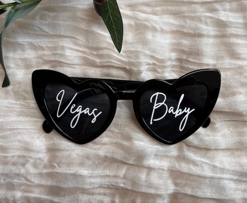 May include: Black heart-shaped sunglasses with white text that reads "Vegas" on the left lens and "Baby" on the right lens.