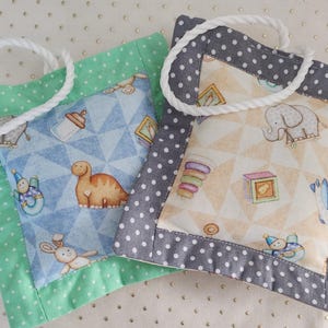 May include: Two handmade baby gift bags. One is mint green with white polka dots, the other is gray with white polka dots. Both have a white rope handle and a central square with baby-themed patterns, including dinosaurs and elephants.