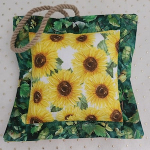 May include: A square fabric bag with a rope handle. The center features a pattern of yellow sunflowers on a white background. The border is green with a leaf pattern.